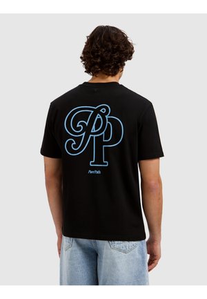 Mann mit lockigem Haar trägt ein schwarzes T-Shirt mit großem blauem "PP" und dem Text "Pure Path" auf dem Rücken, kombiniert mit hellblauen Jeans.