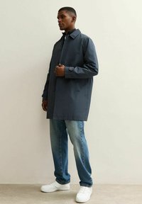 Gabardina gris oscuro con cuello, cierre de botones y mangas largas, combinada con jeans azul claro y zapatillas blancas. Textura suave, diseño minimalista.