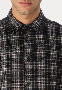 Homme portant une chemise à carreaux noir et blanc à boutons avec col, visible du cou jusqu'au milieu de la poitrine, sur un fond blanc uni.