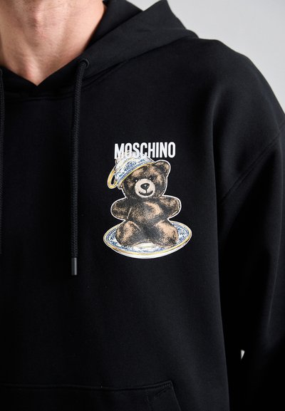 Schwarzer Hoodie mit einem Grafikdesign eines braunen Teddybären auf einem dekorativen Teller, mit dem Text "Moschino" über dem Design. Weiches Baumwollmaterial.
