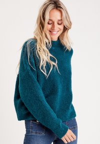 Suéter esponjoso color verde azulado con un cuello alto y corte holgado. Presenta mangas largas y una superficie texturizada. Se combina con jeans de mezclilla oscura.