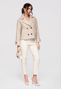 Femme en manteau court croisé beige, pantalon blanc sur mesure, talons nude pointus, tenant un petit sac à main blanc, debout devant un fond clair uni.