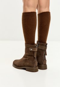Piernas que llevan calcetas de canalé marrón oscuro hasta la rodilla y botas de ante marrón con cremalleras en la parte posterior y detalles de correa sobre un fondo claro.