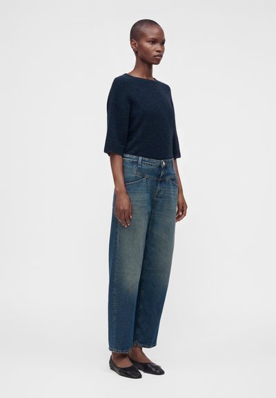 Femme aux cheveux courts portant un haut bleu foncé à manches trois-quarts, un jean bleu ample et des chaussures plates noires, debout devant un fond uni.