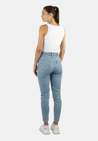 Woman with brown hair in a ponytail wearing gold earrings, white sleeveless top, light blue high-waisted jeans, and white sneakers, facing back.