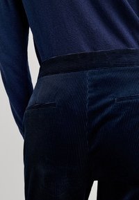 Granatowe spodnie z korduroju o żebrowej fakturze, z dwoma tylnymi kieszeniami i gładkim pasem, prezentowane na neutralnym tle.