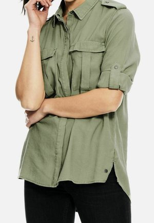 Olive green, short-sleeve shirt with button-up front, two chest pockets, and rolled sleeves. Soft fabric with a slightly loose fit and rounded hem.