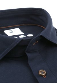 Donker marineblauwe overhemdkraag met bruine steken bij de knoopsgat, een bruine knop en een wit label aan de binnenkant met een gestileerde "R".
