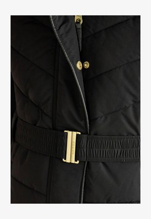 Detailný záber čiernej prešívanej bundy so zlatými cvokmi, zipsom a čiernym elastickým pásom so zlatou prackou s vyrytým nápisom "JOULES."