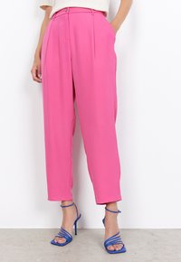 Pantalons roses ajustés avec une texture lisse, des plis à l'avant et une taille haute. Associés à des talons hauts strappy bleus.