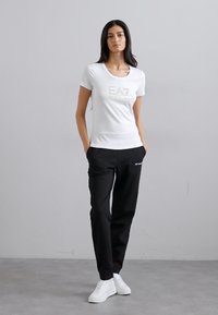 T-shirt bianca con logo argento "EA7", abbinata a pantaloni neri con dettagli del logo e sneakers bianche. Abbigliamento sportivo semplice e casual.