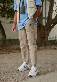 Beiga strukturerade byxor med en liten svart Nike-logotyp, kombinerade med en ljusblå skjorta och mönstrade vita sneakers med gröna och orange detaljer.