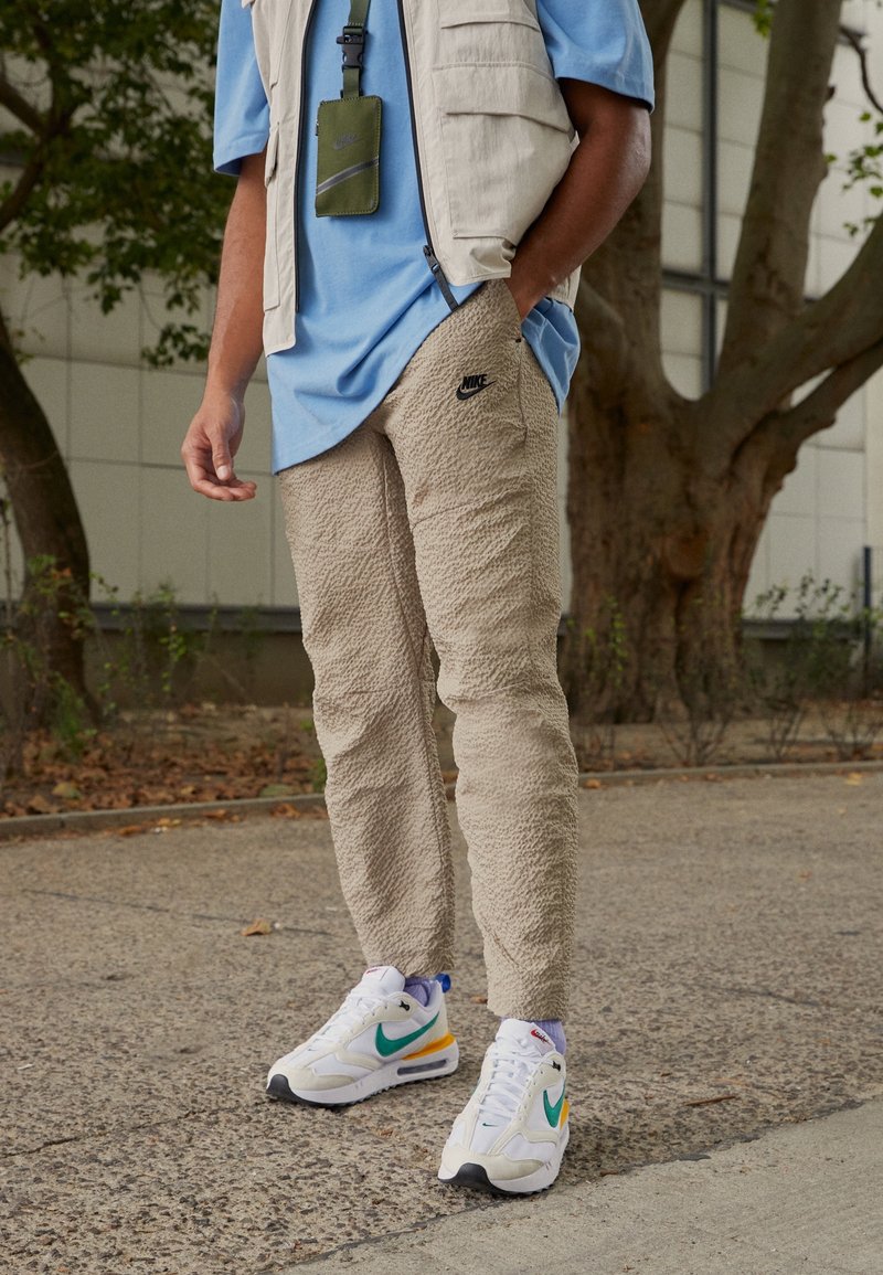 Beiga strukturerade byxor med en liten svart Nike-logotyp, kombinerade med en ljusblå skjorta och mönstrade vita sneakers med gröna och orange detaljer.