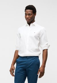 Άνδρας που φοράει λευκό πουκάμισο με κουμπιά και σηκωμένα μανίκια και μπλε παντελόνι, στέκεται μπροστά σε απλό φωτεινό φόντο, κοιτάζοντας προς τα πλάγια.