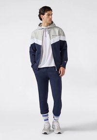 Marineblauw en grijs sportief trainingspak met een ritssluiting in de hoodie, witte accenten en een losse pasvorm. Inclusief gestreepte sokken en lichtgrijze sneakers.