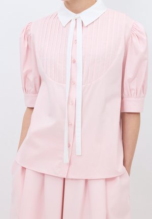 Femme portant un chemisier rose clair avec un col blanc, des plis verticaux sur la poitrine, des manches courtes bouffantes et un nœud blanc au cou.