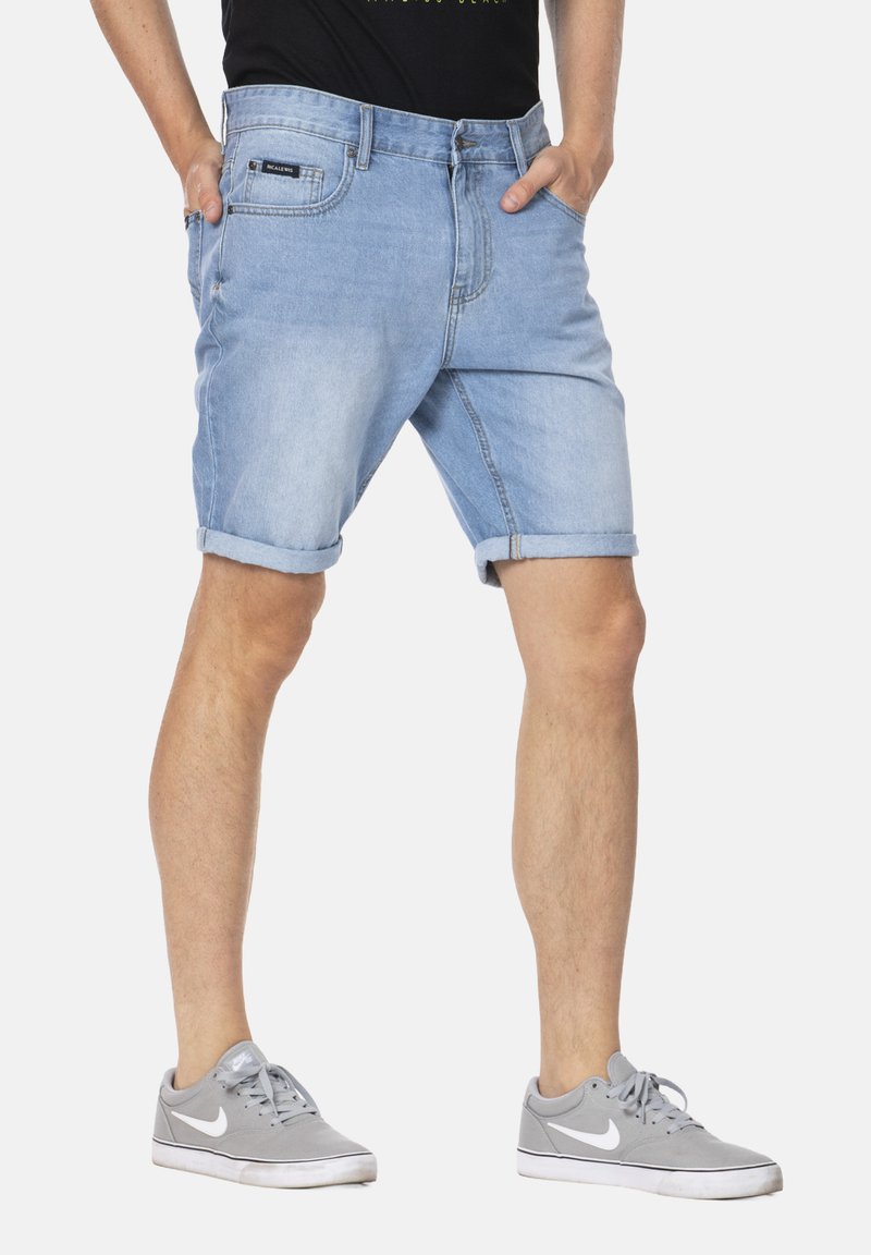 Shorts en denim bleu clair avec une coupe décontractée, ourlets retroussés, deux poches avant et un zip, portés avec des baskets grises.
