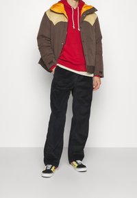 Homme portant une veste marron et jaune par-dessus un sweat à capuche rouge, un pantalon en velours côtelé noir et des baskets noires avec des flammes jaunes et rouges, debout contre un mur blanc.