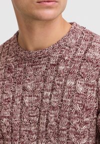 Pull en maille bordeaux avec un col rond, présentant un motif en torsades et des fils contrastants de couleur crème claire, avec des détails de surface texturés visibles.