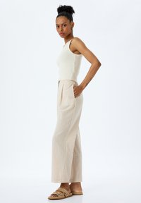 Top de tirantes beige con canalé combinado con pantalones de raya ancha en beige claro. El modelo lleva sandalias color arena con tiras y tiene un peinado natural.