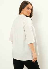 Hellgraues, locker sitzendes Oberteil mit umgeschlagenem Kragen und Dreiviertelärmel. Verfügt über einen Knopfdetail an den Ärmeln. Weicher Stoff.