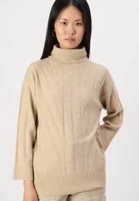 Bežs turtlenecks džemperis, izgatavots no mīkstas auduma; ar vertikālu ribotu rakstu, brīvu piegriezumu un ribotām piedurknēm ar apakšmalu.