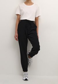 Czarne spodnie dresowe z elastycznym pasem i sznurkiem, bocznymi kieszeniami oraz ściągaczami na nogawkach, zestawione z białą koszulką z krótkim rękawem i szarymi trampkami.