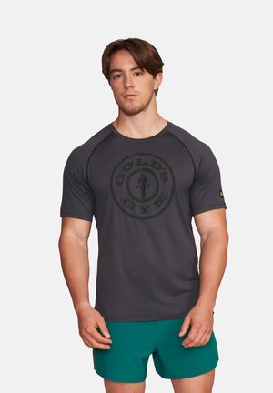 Junger Mann mit braunen Haaren, der ein eng anliegendes dunkelgraues Gold's Gym T-Shirt und grüne Sportshorts trägt und vor einem schlichten Hintergrund steht.
