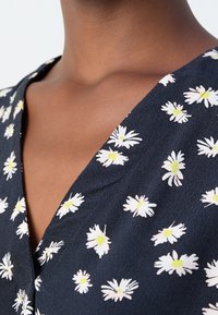 Gros plan sur le cou et le haut de la poitrine portant un vêtement bleu marine avec un motif floral de marguerites blanches et jaunes et un décolleté en V.