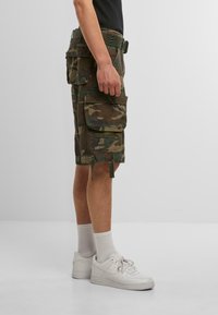 Tarnfarben Cargo-Shorts in Brauntönen und Grüntönen mit mehreren Taschen, verstellbarem Saum und elastischem Bund. Getragen mit weißen Sneakers.