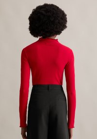 Rotes, geripptes Langarmoberteil mit einem rüschierten hohen Kragen, kombiniert mit hoch taillierten schwarzen Hosen, die eine dezente Nadelstreifenstruktur aufweisen.