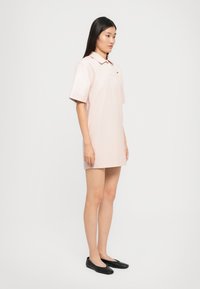 Rochie polo dintr-un material roz deschis, cu guler clasic, mâneci scurte și croială dreaptă. Purtați cu balerini negri.
