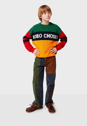 Strickpullover mit grünen, roten und orangefarbenen Abschnitten, mit "BOBO CHOSES"-Text. Patchwork-Jeans in grün, braun, blau und dunkelblau. Braune Schuhe.