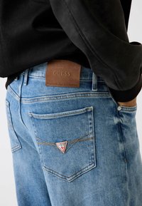 Dżinsy w jasnoniebieskim kolorze z paskiem na talii z naszywką z logo GUESS ze skóry. Cechy charakterystyczne to beżowe szwy i trójkątne logo na tylniej kieszeni.