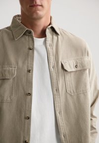 Camicia in corduroy beige con due tasche sul petto, chiusura a bottoni e colletto alla francese, indossata sopra una maglietta bianca. Tessuto testurizzato con righe visibili.