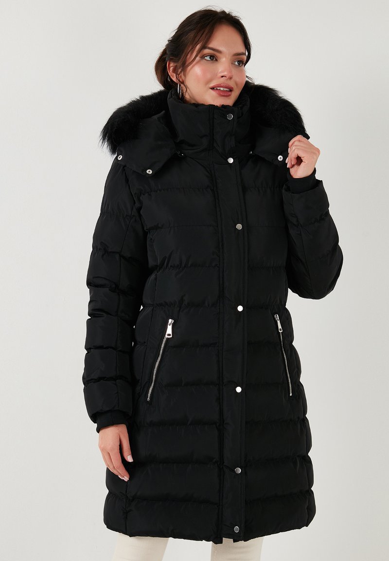 Sort pufferjakke med faux fur-fór i hætten, sølvfarvede lynlåse og en quiltet design. Mellemlang med lange ærmer og en frontlås med trykknap.