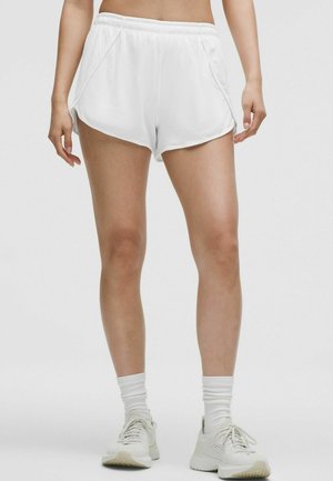 Gambe che indossano pantaloncini sportivi bianchi, calzini bianchi alla caviglia e scarpe da corsa bianche, in piedi su uno sfondo grigio chiaro uniforme.