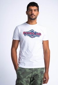 Camiseta blanca de algodón con un estampado gráfico en rojo y azul que dice "PETROLIND" junto con texto adicional; combinada con pantalones cortos cargo de camuflaje verde.