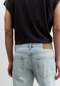 Helle blaue Denim-Jeans mit einem rechteckigen Lederpatch. Die Gesäßtaschen sind in einer spitzen Form genäht. Schwarzes ärmelloses Oberteil sichtbar.