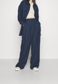 Marinblå oversized skjorta, beige croppat top och vida marinblå byxor med veck. Vita sneakers fulländar outfiten. Slät tygtextur.