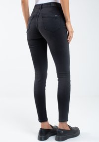 Vaqueros negros ajustados con una cintura alta, diseño de cinco bolsillos y detalles de costura sutiles. Llevados con zapatos negros tipo slip-on.