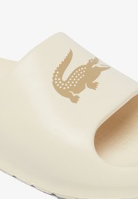 Beige rubber slide sandal with an open toe, featuring a crocodile logo in a darker beige shade on the upper section. Textured sole.
