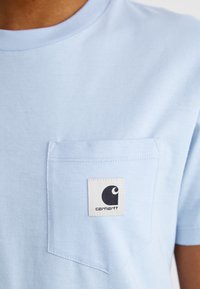 Svijetloplava pamučna majica s okruglim izrezom i džepom na prsima s crno-bijelim oznakom Carhartt. Glatka tekstura.