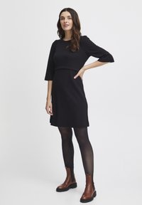 Svart knälång klänning med trekvartsärmar, texturerat material och en figurnära överdel. Modellen bär svarta strumpbyxor och bruna ankelkängor.