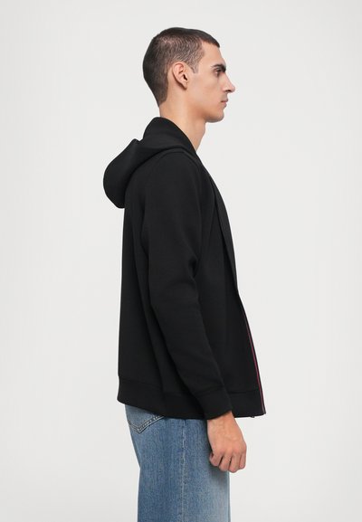 Čierna mikina na zips s kapucňou, vyrobená z mäkkej tkaniny. Obsahuje predný zips a rebrované manžety, skombinovaná s svetlými denim jeans.