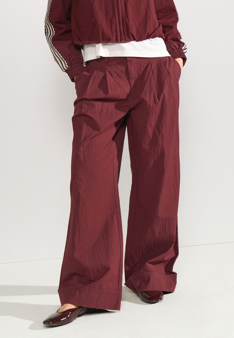 Pantalon ample rouge foncé avec un motif à carreaux subtil, porté avec une veste assortie et des chaussures plates rouge foncé.