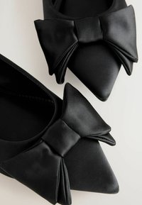 Ballerines noires en satin avec un bout pointu et un grand nœud sur le devant. Texture lisse avec un léger éclat.