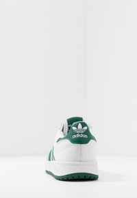 White sneaker with green accents, featuring horizontal ribbed texture, three green stripes, and a rubber sole. Adidas logo on heel.