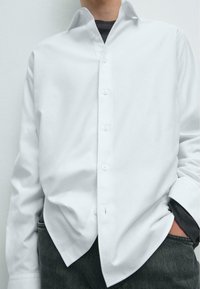 Weißes Hemd mit Button-Down-Kragen, aus glattem Stoff, mit lockerer Passform und tief eingesetztem Schulterdesign. Getragen über dunklen Jeans.