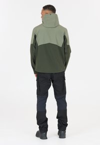 Veste verte et vert foncé, avec un col montant et une capuche. Associée à un pantalon cargo noir avec des panneaux aux genoux et des poches.